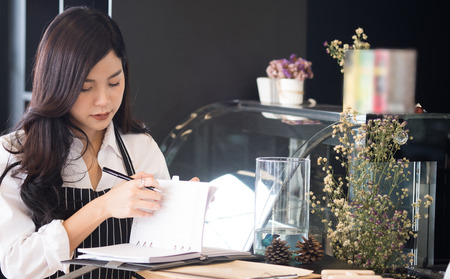 small business owner with notebook at counter in coffee shop. asian female barista wearing apron at bar in cafe. food service, restaurant, entrepreneur concept.の写真素材