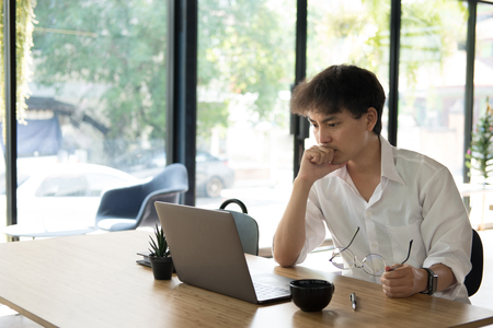asian student using computer for learning leasson online at cafe. startup man working with laptop at office.  young male entrepreneur analyze financial data at workplace. business, education conceptの写真素材