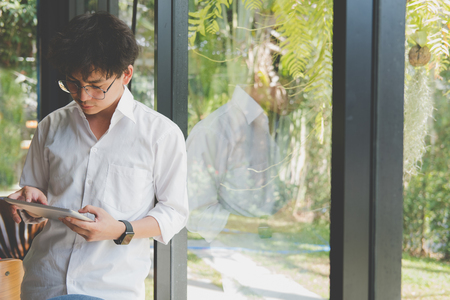 asian businessman using tablet computer at workplace. startup man working with touchpad at office.  young male entrepreneur holding pad standing at window in cafeの写真素材