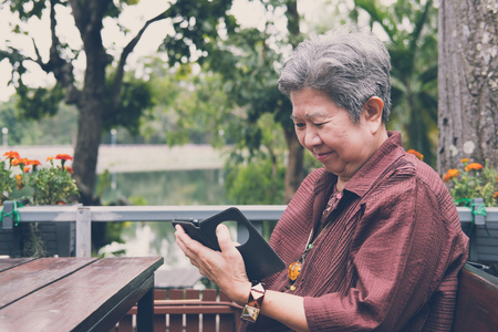 Asian elder woman holding mobile phone in garden. elderly female texting message, using app with smartphone in park. senior use cellphone to connect with social networkの写真素材