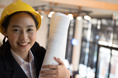 asian female engineer holding construction blueprint. young architect wearing yellow plastic helmet hardhat at workplaceの写真素材