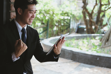 Successful business man checking tablet message & raise fist with happiness and gladness outside office building. male startup entrepreneur enjoy success in job. communicate technology in business achievement conceptの写真素材