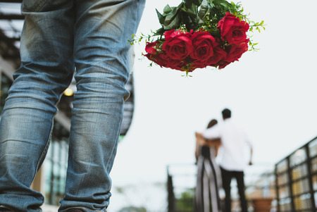 asian heartbroken man holding bouquet of red roses feeling sad while seeing woman dating with another man. broken heart, disappointed in love conceptの写真素材