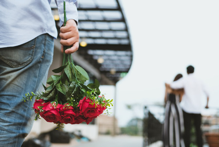 asian heartbroken man holding bouquet of red roses feeling sad while seeing woman dating with another man. broken heart, disappointed in love conceptの写真素材