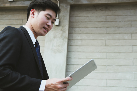 confident businessman in suit holding touchpad while standing outside building. young asian man using digital tablet for work outdoorsの写真素材