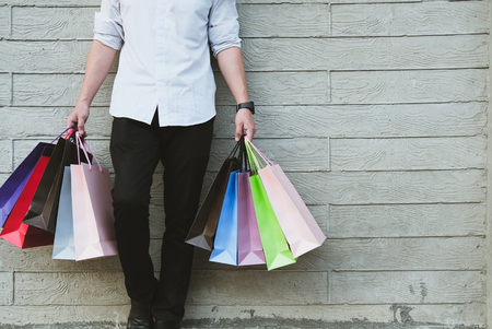 young man holding colorful shopping bag outdoors. shopaholic male standing beside gray wall. shopper enjoy buying. consumerism conceptの写真素材