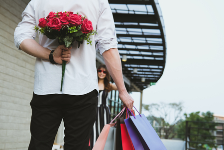 man holding shopping bag & bouquet of red roses behind his back for woman. boyfriend surprise girlfriend with flower in valentine's day. celebrate anniversary, love & relationship conceptの写真素材