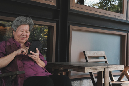 asian elder woman holding mobile phone on terrace. elderly female texting message, using app with smartphone on patio. senior use cellphone to connect with social networkの写真素材