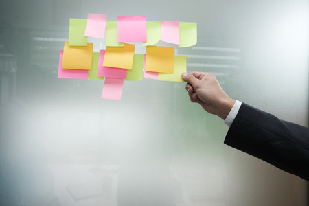 Businessman hold adhesive notes on glass wall at workplace. Sticky note paper reminder schedule for discussing creative idea & business brainstormingの写真素材