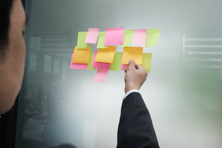 Businessman hold adhesive notes on glass wall at workplace. Sticky note paper reminder schedule for discussing creative idea & business brainstormingの写真素材
