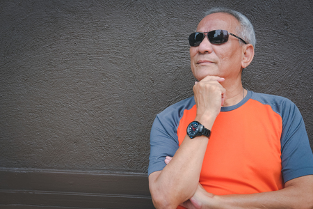 thoughtful elder man wearing sunglasses standing near brown wall. asian elderly male thinking. senior leisure lifestyleの写真素材