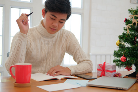 man wearing cream sweater writing note on notebook at home during christmas new year holiday celebration.の写真素材