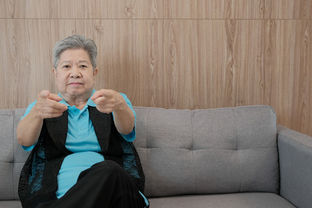 old asian elderly woman smiling pointing at camera. happy elder senior sit on sofa at homeの写真素材