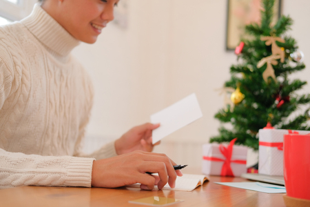 man wearing cream sweater writing christmas letter greeting card holiday wishes with xmas decoration.の写真素材
