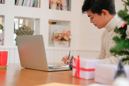 man wearing cream sweater writing christmas letter greeting card holiday wishes with xmas decoration.の写真素材