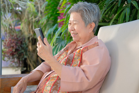 asian elder woman holding mobile phone on terrace. elderly female using smartphone on patio. senior texting message with cellphoneの写真素材