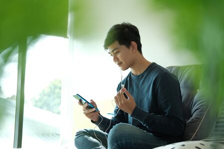 asian male teenager man listening to music from smart mobile phone while sitting on floor at homeの写真素材