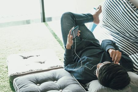 asian male teenager man listening to music from smart mobile phone while lying on floor at homeの写真素材