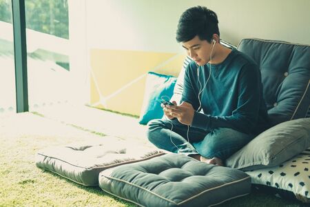 asian male teenager man listening to music from smart mobile phone while sitting on floor at homeの写真素材
