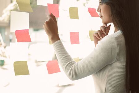 businesswoman woman thinking planning with adhesive notes on glass wall at workplace. Sticky note paper reminder schedule for creative idea & business brainstormingの写真素材