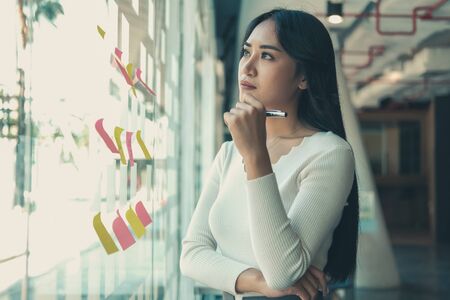 businesswoman woman thinking planning with adhesive notes on glass wall at workplace. Sticky note paper reminder schedule for creative idea & business brainstormingの写真素材