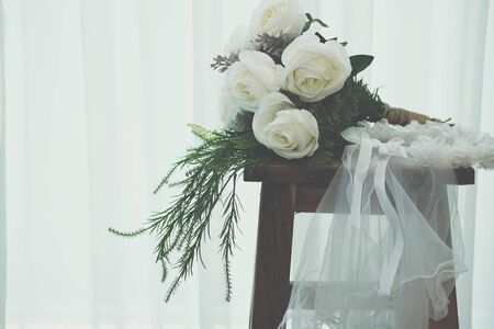 wedding white bridal veil & rose flower bouquet on wooden chair beside see through sheer window curtainの写真素材