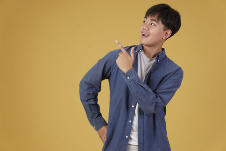 portrait of happy young asian man dressed casually pointing finger up at copyspace. yellow studio...