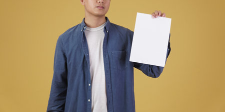 portrait of young asian man dressed casually showing blank empty placard paper isolated on yellow studio background.の写真素材