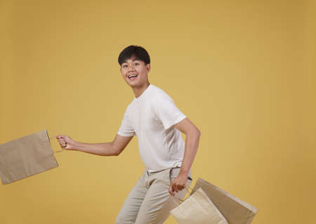 portrait of happy young asian man dressed casually holding shopping bags isolated on yellow studio backgroundの写真素材