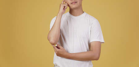 portrait of thoughtful young asian man dressed casually thinking doubting wondering isolated on yellow studio backgroundの写真素材