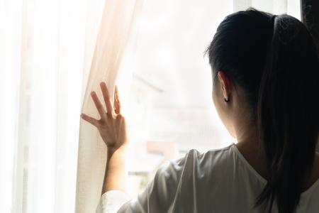 sad woman holding the curtains open to look out of a large light window at home, interiorの写真素材