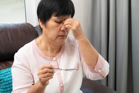 Tired old woman removing eyeglasses, massaging eyes after reading paper book. feeling discomfort because of long wearing glasses, suffering from eyes pain or headacheの写真素材