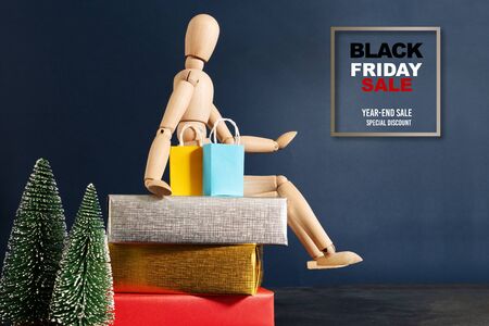 Black Friday sale, wooden doll sitting on gift box with shopping bag and christmas treeの写真素材