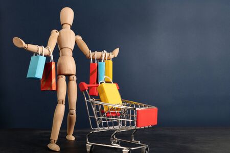 Cheerful happy shopaholic wooden doll with lots of shopping bags on arm and shopping cartの写真素材