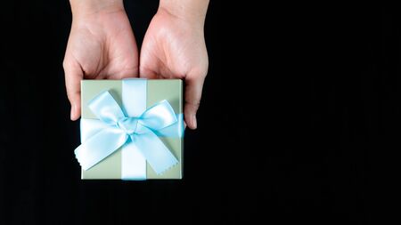 female hands holding a small gift wrapped with blue ribbon on black backgroundの写真素材