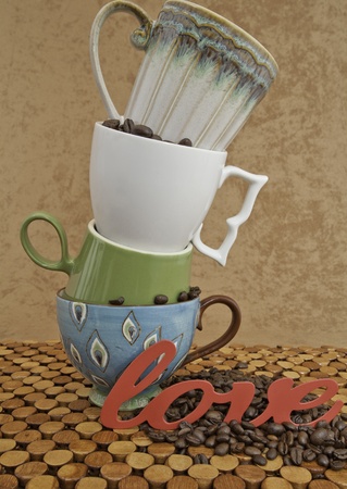 Four Cups Piled on Top of Each Other. Colorful coffee cups piled on each other with coffee beans on wooden mat and in the cups. The word love ha been placed in front of the cups with brown stucco backgound.  の写真素材