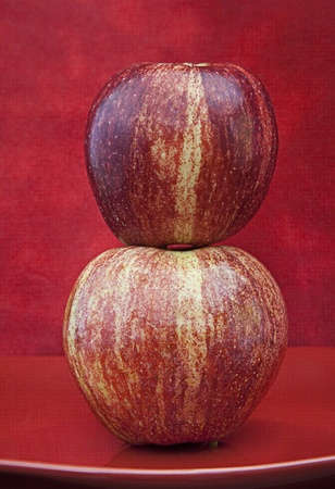 Two Red Apples on top of each other with yellow stripes lined up sitting on a red plate against a textured red background.の写真素材