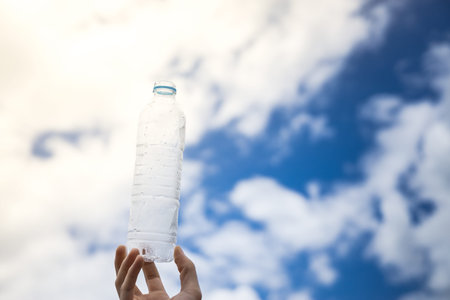 Man holding empty transparent plastic drinking water bottle without cap against the sky in cloudy...