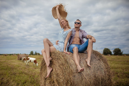 Couple sits on haystack in village against a backdrop of cows, girl raised straw hat up