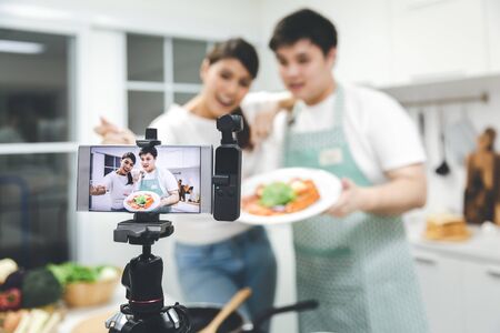Vlogger and blogger freelance job concept, Young asian couple cooking together and recording live video for her vlog and social media with mobile phone camera and tripodの写真素材