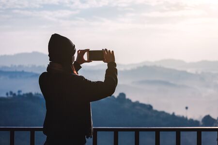 Young travel asian woman with sweater, mobile phone and Wool hat taking photo view of mountain and fog at morning in Khao Kho mountain, Phetchabun, Thailand, famous place in winter seasonの写真素材