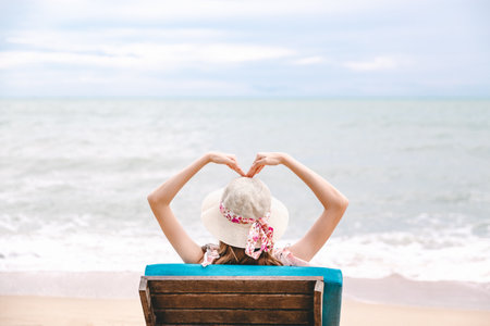 Summer travel beach vacation concept, Happy traveler asian woman with hat relax and making heart shape on chair beach at Pattaya, Chon Buri, Thailandの写真素材