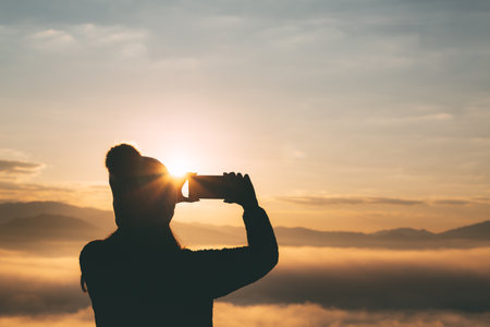 Winter travel relax vacation concept, Young traveler asian woman with sweater and wool hat taking photo and selfie with mobile phone on mountain with fog at sunrise in Mae Hong Son, Thailandの写真素材