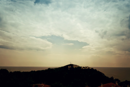 A silhouette of a holy christian cross illuminated by sunlight on top of a hill on a dramatic cloudy sky. Beautiful view at the coast of the Black Sea in Balchik city, Bulgaria.の写真素材