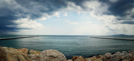 Sea landscape between two piers on the stone beach with a dramatic cloudy sky. Beautiful view at the coast of the Black Sea in Balchik city, Bulgaria.の写真素材