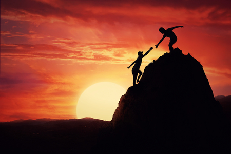 Hikers climbing on rock, mountain at sunset, one of them giving hand and helping to climb. Friendly assistance and support  in a dangerous situation.の写真素材