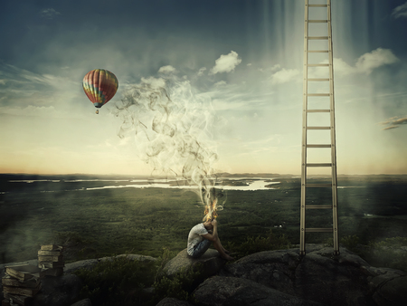 Young boy sit on the top of a hill near a ladder goind up in the sky and a hot air balloon fly over the forest valley. Bad mood, hard thinking, no inspiration concept with his head fired and the smoke rising up.の写真素材