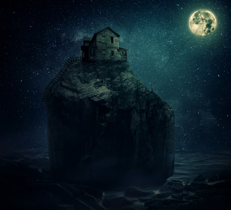 Surreal image with a wood house on the top of a rock hill, near the sea, beyond the starry night sky and a young man, holding a rope, try to catch and pull the full moon.の写真素材