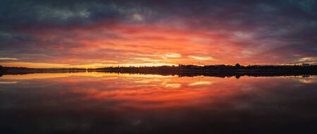 Bold sunset panorama over Delia lake, Moldova. Cloudy dusk sky and red sunlight reflection on the water surface. Dramatic dusk waterscape.の写真素材