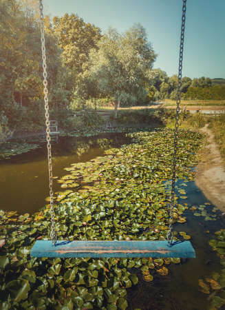 Vertical photo of a vintage blue swing hanging over a pond with blooming waterlily, lotus flowers. Vacation travel, natural background at the lake recreation concept, at Bardar village, Moldova.の写真素材